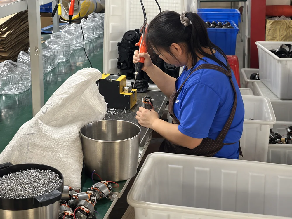 Worker using an orange assembly tool beside motors, glass bowls and component bins